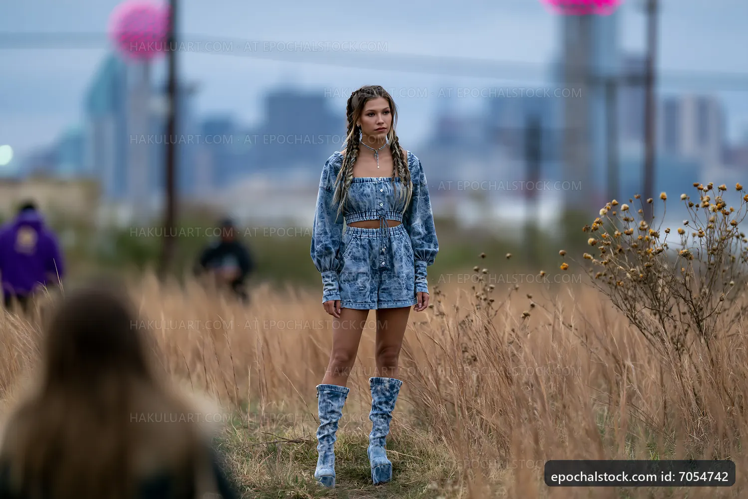 Young woman in denim outfit stands in tall grass with city skyline