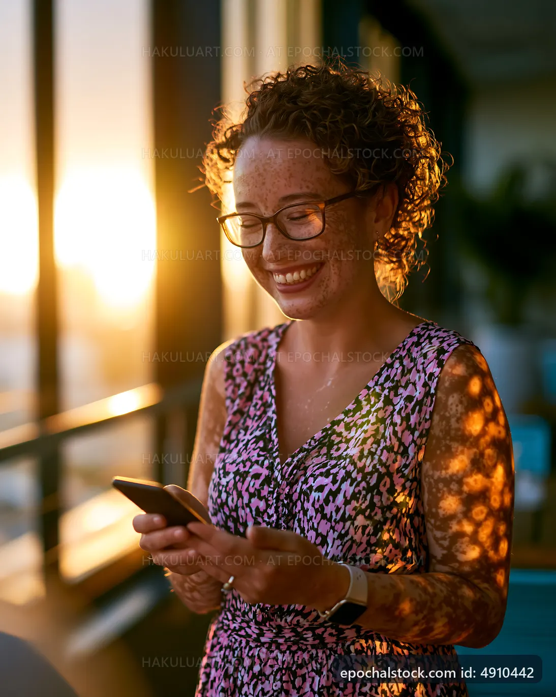 Woman smiles while using a smartphone near a sunset window