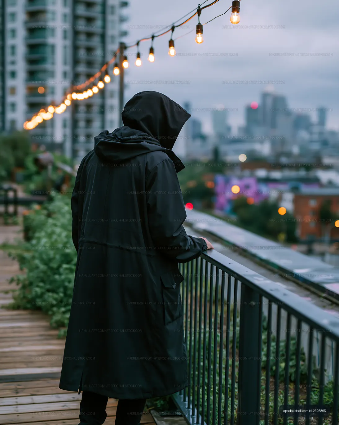 Person in black raincoat overlooking city with string lights